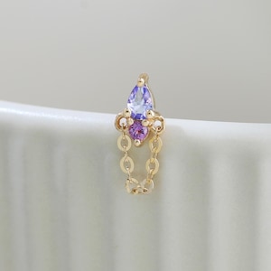 May include: A gold stud earring featuring a marquise-cut light blue gemstone positioned above a round purple gemstone, with a delicate gold chain loop hanging from the base of the setting, creating a vertical drop design for the ear.