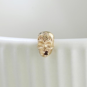 May include: A small gold-toned stud earring shaped like a skull with intricate filigree-style engravings on the forehead and cheekbones, featuring a dark, recessed eye socket detail on one side, resting against a white ribbed surface.