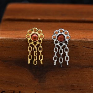 Könnte beinhalten: Zwei blumenförmige Ohrstecker, einer goldfarben und einer silberfarben, jeweils mit einem rotbraunen Stein in der Mitte und drei hängenden Kettengliedern. Die Ohrringe sind auf einem Holzblock mit der Aufschrift GoldenAuraPiercings platziert.