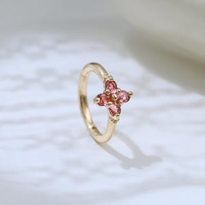 May include: A gold hoop earring featuring a small, four-petaled flower charm set with four pink gemstones. The delicate metal band is circular, and the floral accent is positioned at the top of the hoop, creating a refined and simple jewellery piece.