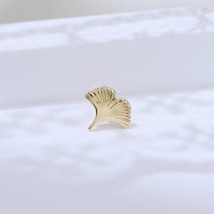 May include: A small, gold-colored ginkgo leaf-shaped stud earring. The earring has a detailed, textured surface and a curved, fan-like design. The earring is set against a white background.