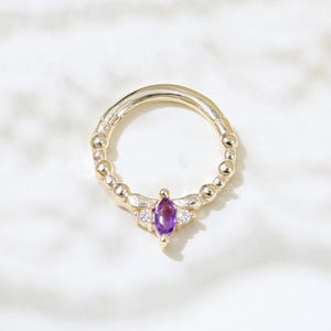 May include: A gold ring featuring a central, marquise-cut purple gemstone, with two smaller white gemstones. The ring band has a beaded design. The ring is set against a white background.