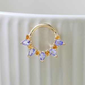 Könnte beinhalten: Ein goldfarbener Septumring mit abwechselnd blauen und orangefarbenen Edelsteinen. Die tropfenförmigen Edelsteine sind in einem Halbkreis angeordnet. Der Ring befindet sich vor einem weißen Hintergrund. Die Steine sind ca. 0,5 cm lang.