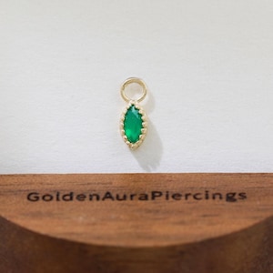 May include: A gold earring charm featuring a teardrop-shaped, emerald-green gemstone. The charm is displayed on a wooden stand with the text "Golden Aura Piercings". The background is white.