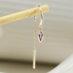 Puede incluir: Un solo pendiente de color dorado con una pequeña gema púrpura en forma de diamante y una barra colgante. El pendiente está suspendido de un fino gancho curvado. El pendiente está sujeto por un palo de madera.
