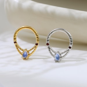 May include: Two hoop earrings, one gold and one silver, each featuring a teardrop-shaped blue gemstone pendant. The gold earring has red accents, while the silver earring has similar red accents. The earrings are displayed on a white surface.