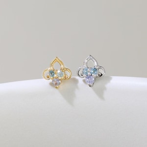 以下が含まれることがあります： 2つのスタッドピアス。1つは金色、もう1つは銀色で、それぞれ4枚の花びらのデザイン。金色のイヤリングには水色と紫色の宝石があり、銀色のイヤリングにも水色と紫色の宝石があります。白い表面にイヤリングが置かれています。