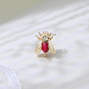 Może przedstawiać: Złota broszka w kształcie pszczoły z rubinowym korpusem i diamentowymi akcentami. Skrzydła są szczegółowe z wytrawionymi liniami. Broszka jest umieszczona na białym tle.