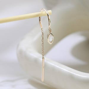Op de afbeelding: Een delicate gouden oorbel met een kleine, heldere edelsteen en een hangende staaf. De oorbel hangt aan een gebogen haak en ketting. De achtergrond is een zachte, neutrale kleur, die het elegante ontwerp van de sieraden benadrukt.
