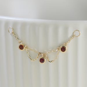 May include: A delicate gold chain connector featuring three small, round, bezel-set dark red gemstones. The piece consists of two thin gold chains draped in a scalloped pattern, with a circular jump ring at each end for attachment to other jewelry.