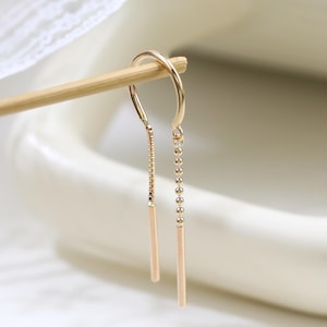 May include: A gold threader earring featuring a delicate chain with a small bead detail on one side and a smooth, straight bar at the end of each strand. The earring is suspended from a thin wooden stick, showcasing its slender, minimalist design.