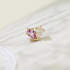 Peut inclure: Une petite boucle d'oreille à clou dorée avec deux pierres précieuses en forme de cœur. L'une est rose, l'autre claire. La boucle d'oreille est posée sur une surface blanche texturée, avec de la dentelle en arrière-plan.