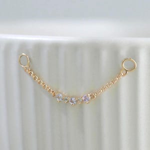 May include: A delicate gold-tone chain connector featuring three small, clear, round-cut stones set in the center. The chain terminates in two small circular jump rings on each end, designed for linking jewelry components together in a curved arrangement.