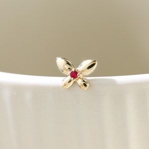 Op de afbeelding: Een klein, goudkleurig sieraad in de vorm van een vlinder met een rode edelsteen in het midden. De vlinder is geplaatst op een wit, gebogen oppervlak, met een zachte, neutrale achtergrond. Het sieraad is waarschijnlijk een oorbel of een kleine hanger.