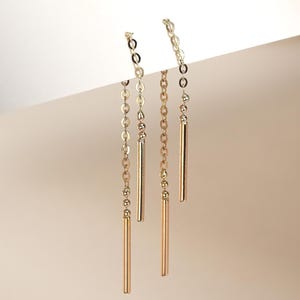 May include: A pair of gold threader earrings featuring a delicate fine chain that suspends a slender, polished gold bar at the end. The earrings are designed to be pulled through the earlobe, allowing the chain and bar to hang vertically.