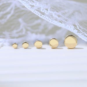 Peut inclure: Une collection de cinq boucles d'oreilles rondes de couleur or, de tailles variées. Les boucles d'oreilles sont disposées en ligne, la plus petite à gauche et la plus grande à droite. Un délicat tissu en dentelle blanche est drapé en arrière-plan.