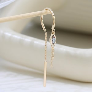 May include: A gold-toned threader earring featuring a curved wire, a small oval-shaped light blue gemstone, and a delicate chain ending in a slender gold-toned bar. The earring is suspended from a thin wooden stick against a soft white background.