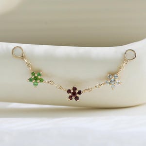 Puede incluir: Un conector de cadena dorada con tres dijes en forma de flor, cada uno engastado con pequeñas piedras preciosas. La flor de la izquierda es verde, la central es de color rojo oscuro y la de la derecha es azul claro. La cadena termina en dos aros dorados.