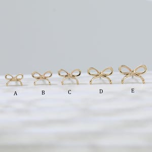 Pode incluir: Uma coleção de brincos de botão dourados em forma de laço. Os brincos estão dispostos em uma fileira, aumentando de tamanho da esquerda para a direita. Os laços têm um design delicado e minimalista. O fundo é de uma cor suave e neutra.