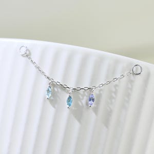 Puede incluir: Una cadena de plata delicada con tres piedras preciosas colgantes. Las piedras preciosas son azul claro, azul oscuro y morado claro.