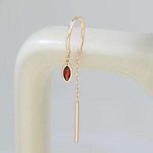 May include: A single gold-colored earring with a dangling chain and a small, oval, red gemstone. The earring is suspended from a curved hook and features a slender, rectangular bar at the bottom.