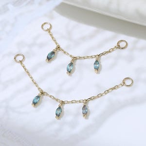 Puede incluir: Dos conectores de joyería en tono dorado con cadenas delicadas y pequeñas piedras preciosas azules de corte marquesa colgando de cada eslabón. Cada pieza termina con un anillo circular en ambos extremos para sujetar a pendientes u otras joyas.