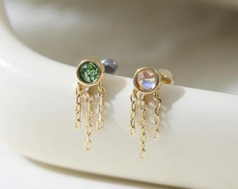May include: Two gold earrings with dangling chains. One earring features a green gemstone, the other a clear gemstone. The earrings are displayed on a white, curved surface. The earrings are small and delicate.