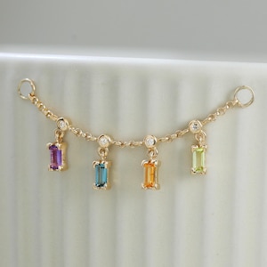 Könnte beinhalten: Zarte goldene Kette mit fünf rechteckigen farbigen Edelsteinen in Lila, Türkis, Orange und Grün. Zwischen jedem farbigen Stein befinden sich kleine, klare Edelsteine. Die Kette ist vor einem weißen Hintergrund abgebildet.