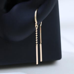 Könnte beinhalten: Ein Paar goldfarbene Ohrhänger. Ein Ohrring hat eine Kette aus kleinen Perlen, der andere eine filigrane Kette. Beide haben am Ende einen geraden, rechteckigen Stab. Die Ohrringe werden vor einem schwarzen Hintergrund gezeigt.