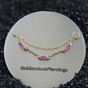 Puede incluir: Un conector de piercing de cadena dorada con dos cadenas delicadamente drapeadas y tres pequeñas piedras preciosas rectangulares de color púrpura. La pieza incluye dos anillos circulares en los extremos y lleva el texto GoldenAuraPiercings.