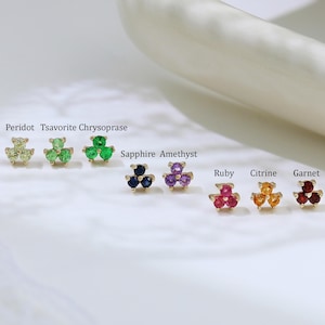 Könnte beinhalten: Goldene Ohrstecker mit jeweils drei Edelsteinen. Zu den Edelsteinen gehören Peridot, Tsavorit, Chrysopras, Saphir, Amethyst, Rubin, Citrin und Granat. Präsentiert auf einer weißen Oberfläche. Maße nicht verfügbar.