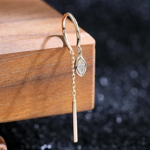 Puede incluir: Un pendiente de hilo de oro con una cadena delicada, una piedra preciosa transparente de talla marquesa en engaste de bisel y una barra de oro delgada en el extremo. El pendiente cuelga de un alambre de oro pulido con un diseño elegante.