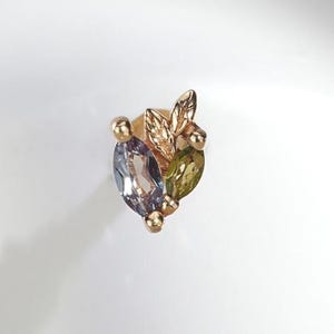Op de afbeelding: Een gouden oorsteker met een clusterontwerp, voorzien van een lichtpaarse edelsteen in marquise-slijpvorm, een olijfgroene edelsteen in marquise-slijpvorm en een klein goudkleurig accent in de vorm van een blaadje.