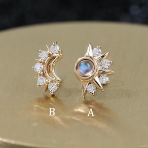 Puede incluir: Un par de pendientes dorados, uno con forma de media luna y otro con diseño de sol. Ambos pendientes están adornados con pequeñas gemas brillantes. Los pendientes se exhiben sobre una superficie dorada, sobre un fondo oscuro.