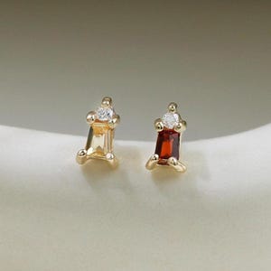 Könnte beinhalten: Ein Paar Ohrstecker mit einer goldfarbenen Metallfassung mit vier Krappen. Ein Ohrring enthält einen hellgelben Edelstein im Baguetteschliff, der andere einen dunkelroten Edelstein im Baguetteschliff, jeweils gekrönt von einem kleinen runden Stein.