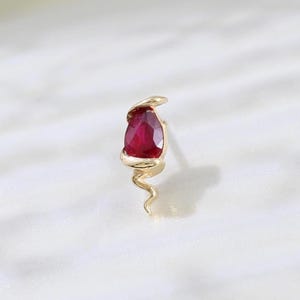 May include: A gold-colored stud earring featuring a teardrop-shaped, deep red gemstone. The earring has a delicate, swirling design below the gemstone. The earring is set against a soft, white background.