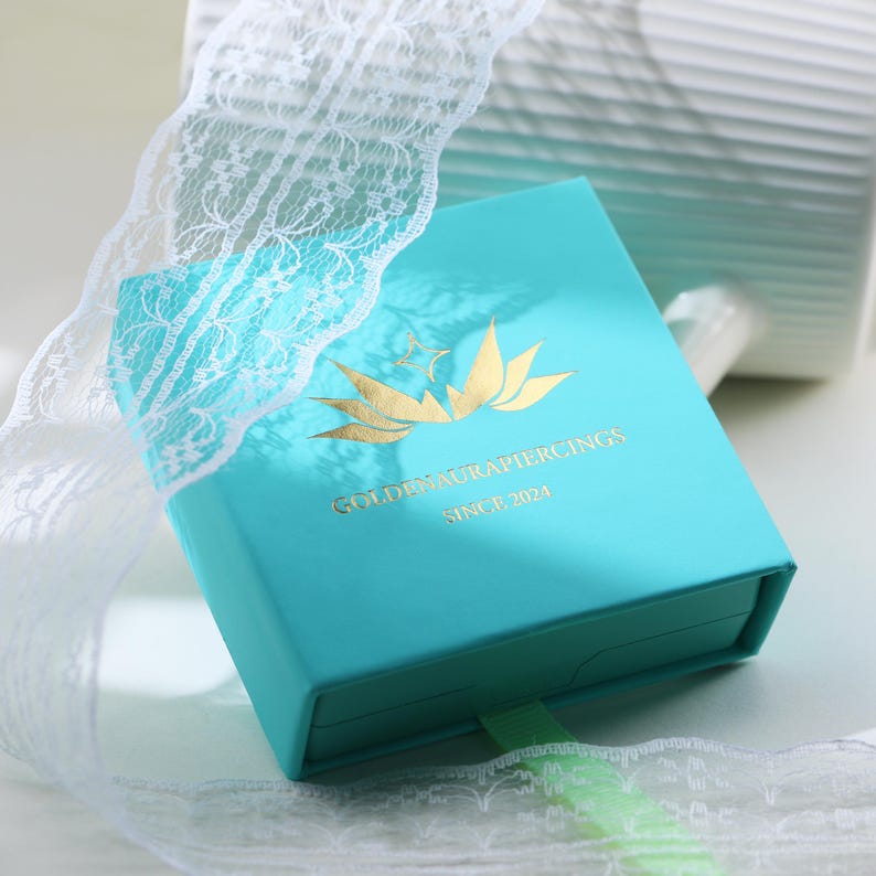 May include: A turquoise jewelry box with gold lettering and a gold logo featuring a bird and floral design. The text on the box reads "GOLDENAURAPIERCINGS SINCE 2024". A white lace ribbon is wrapped around the box.