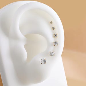 Puede incluir: Un conjunto de seis pendientes de bot&oacute;n dorados con piedras redondas transparentes, dispuestos en una l&iacute;nea vertical a lo largo del borde exterior de un modelo de oreja blanco. Cada pendiente presenta un engaste de cuatro garras.