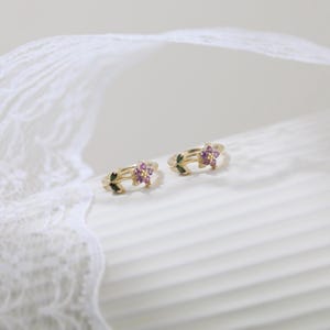 May include: Two gold-colored rings, each featuring a floral design with small, purple gemstones at the center. The rings also have green leaf accents. The background is a soft, neutral color, with a white lace detail.