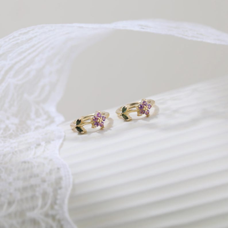 May include: Two gold-colored rings, each featuring a floral design with small, purple gemstones at the center. The rings also have green leaf accents. The background is a soft, neutral color, with a white lace detail.