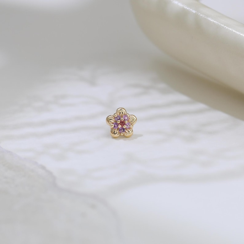 Pode incluir: Um pequeno brinco de ouro em forma de flor de cinco p&eacute;talas. O centro da flor apresenta uma &uacute;nica pedra preciosa vermelha rodeada por cinco pequenas pedras preciosas roxas incrustadas nas p&eacute;talas douradas.