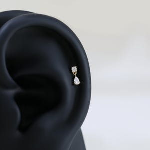 May include: A gold-colored earring with two clear, sparkling cubic zirconia stones. The top stone is square, and the bottom stone is a teardrop shape. The earring is shown on a black ear model.