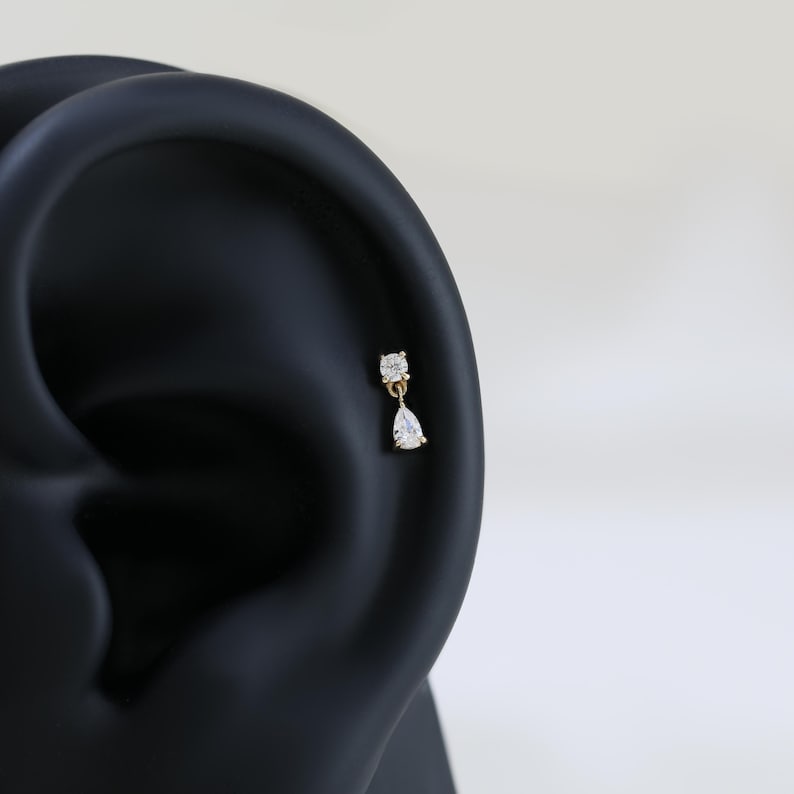 May include: A gold-colored earring with two clear, sparkling cubic zirconia stones. The top stone is square, and the bottom stone is a teardrop shape. The earring is shown on a black ear model.