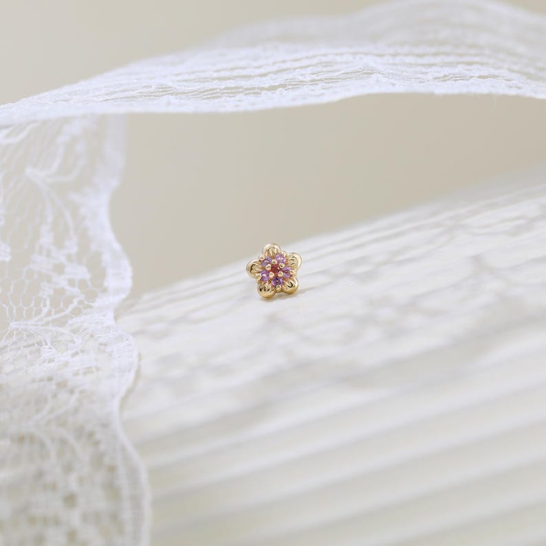 May include: A gold stud earring shaped like a five-petaled flower. The center of the flower features a small red gemstone surrounded by five light purple stones set into each petal, creating a delicate and detailed floral design.