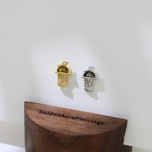 Peut inclure: Deux boucles d'oreilles pendantes en or et en argent avec une pierre pr&eacute;cieuse verte en forme de gobelet &agrave; boba. Les boucles d'oreilles sont pr&eacute;sent&eacute;es sur un support en bois avec le texte "GoldenAuraPiercings".