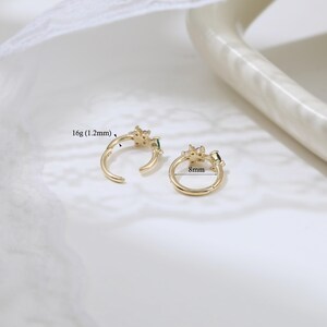 May include: Two gold-colored hoop earrings with a floral design and small green gemstones. One earring is open, and the other is closed. The open earring is labeled "16g (1.2mm)", and the closed earring is labeled "8mm".