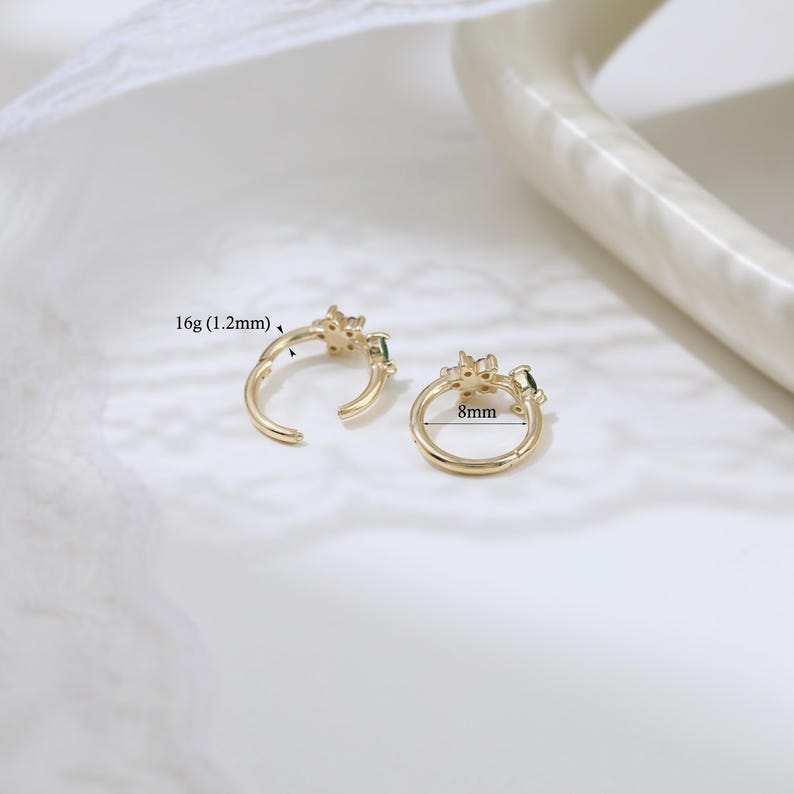 May include: Two gold-colored hoop earrings with a floral design and small green gemstones. One earring is open, and the other is closed. The open earring is labeled "16g (1.2mm)", and the closed earring is labeled "8mm".