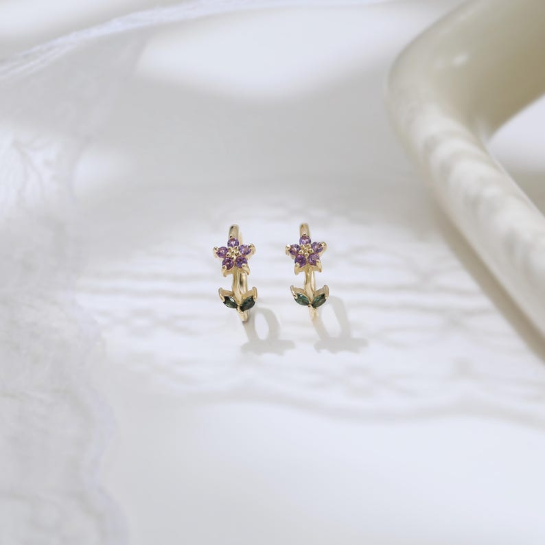 May include: A pair of gold-colored hoop earrings, each adorned with a purple flower design. The flowers are accented with small purple gems and green leaf details. The earrings are set against a white background.