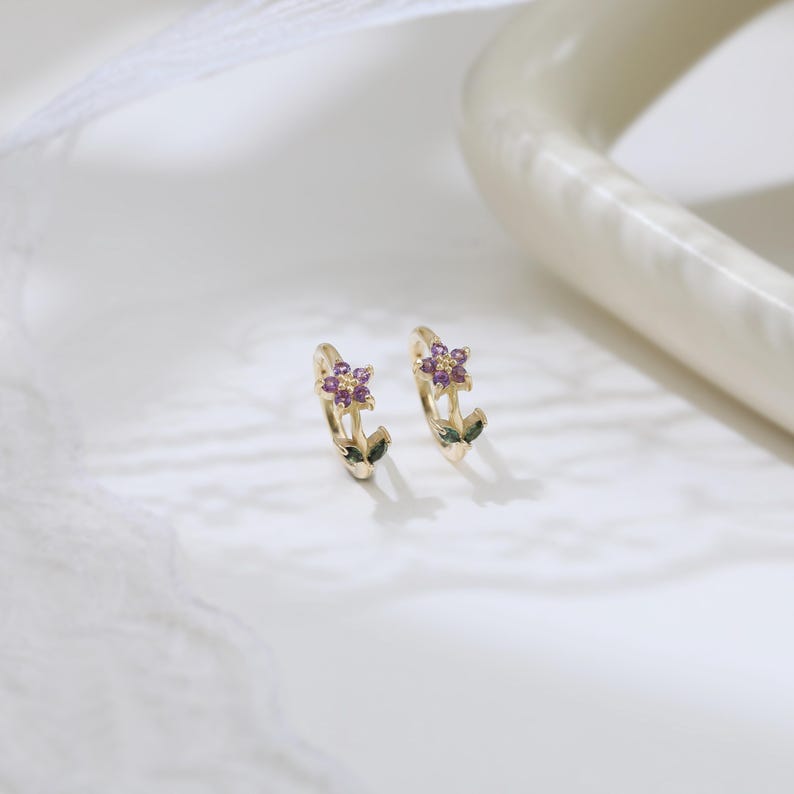 May include: A pair of gold hoop earrings, each adorned with a small purple flower and green leaves. The earrings are set against a white background, highlighting their delicate floral design. The earrings are a stylish accessory.