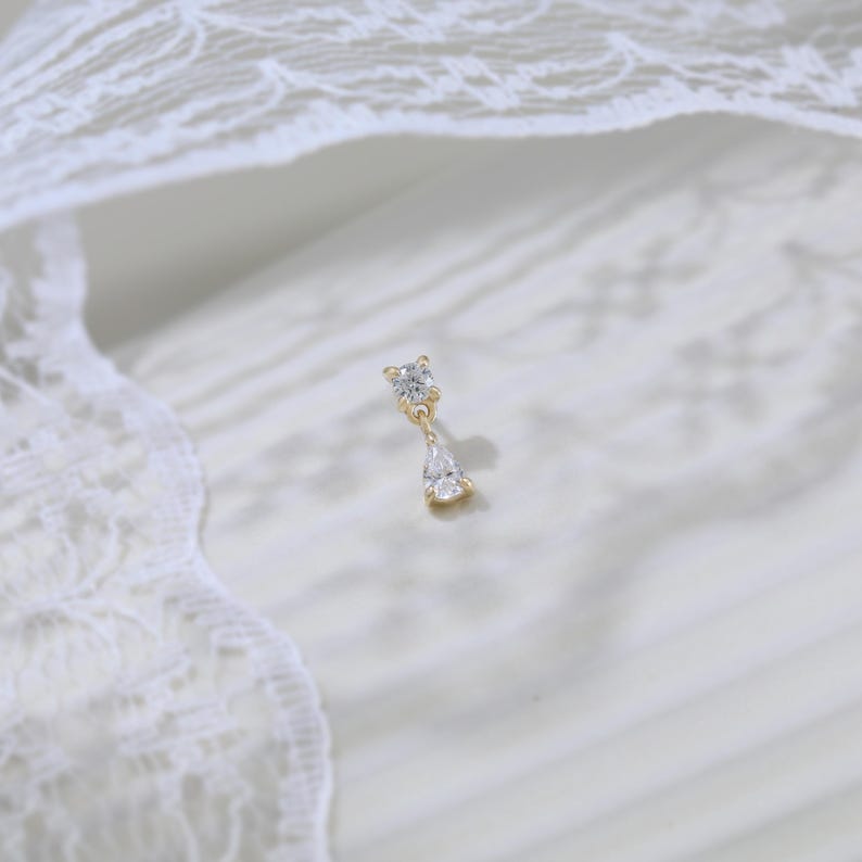 May include: A gold-colored earring with two clear, sparkling gemstones. The top stone is square-shaped, and the bottom stone is a teardrop shape. The earring is set against a white, textured background with lace trim.